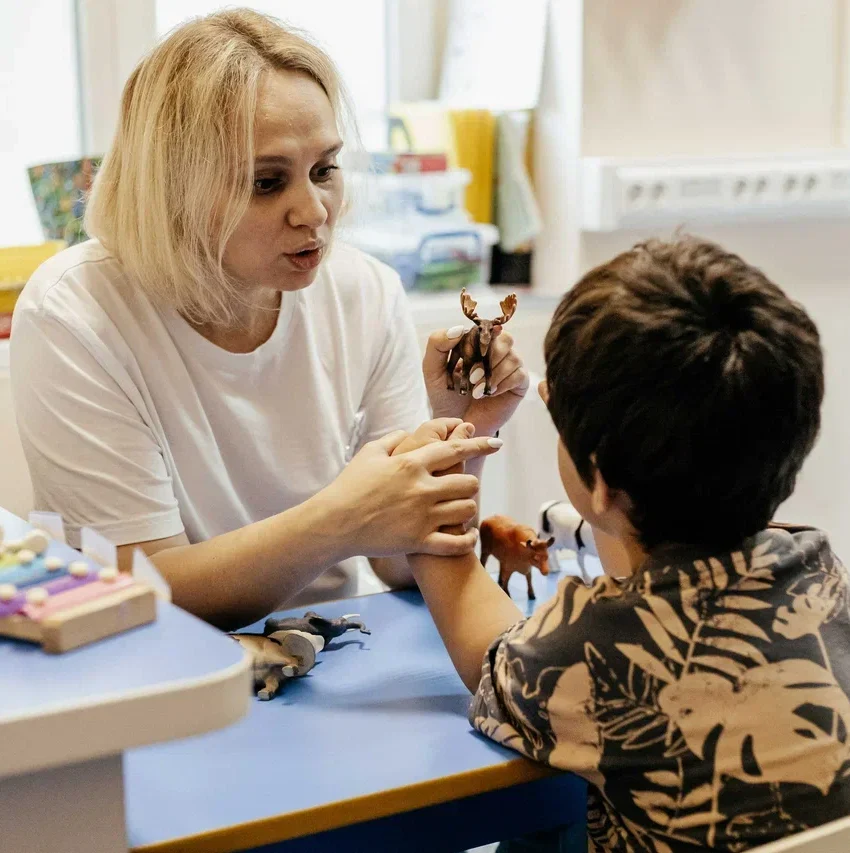 Женщина-специалист в белой футболке сидит за синим столом и проводит коррекционное занятие с мальчиком, держа в руках игрушечных животных, на столе лежат цветные колокольчики и песочные часы.